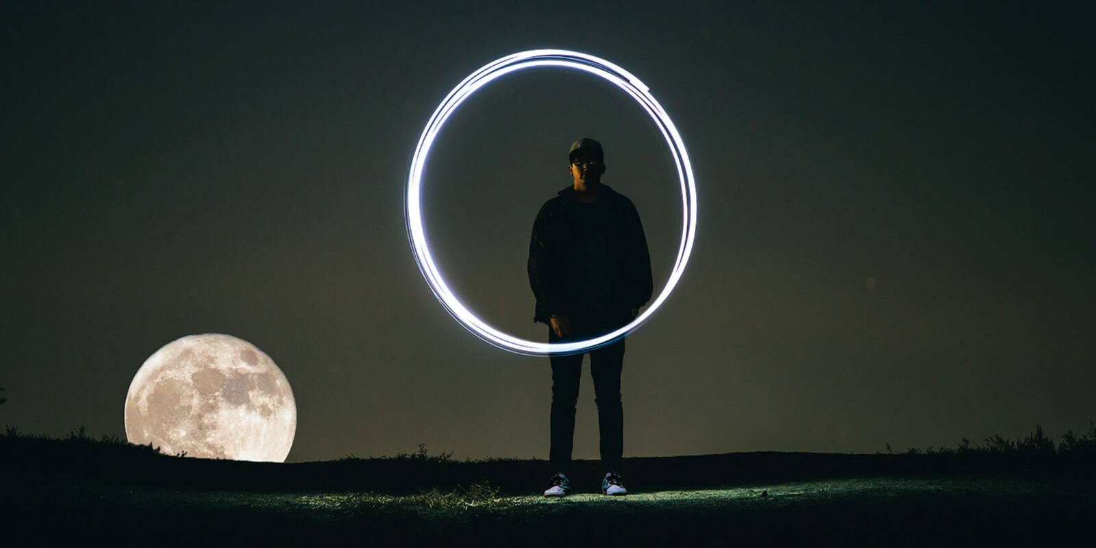 Ein Mann steht in der Nacht auf einer Wiese vor dem Mond und macht einen Lichtkreis